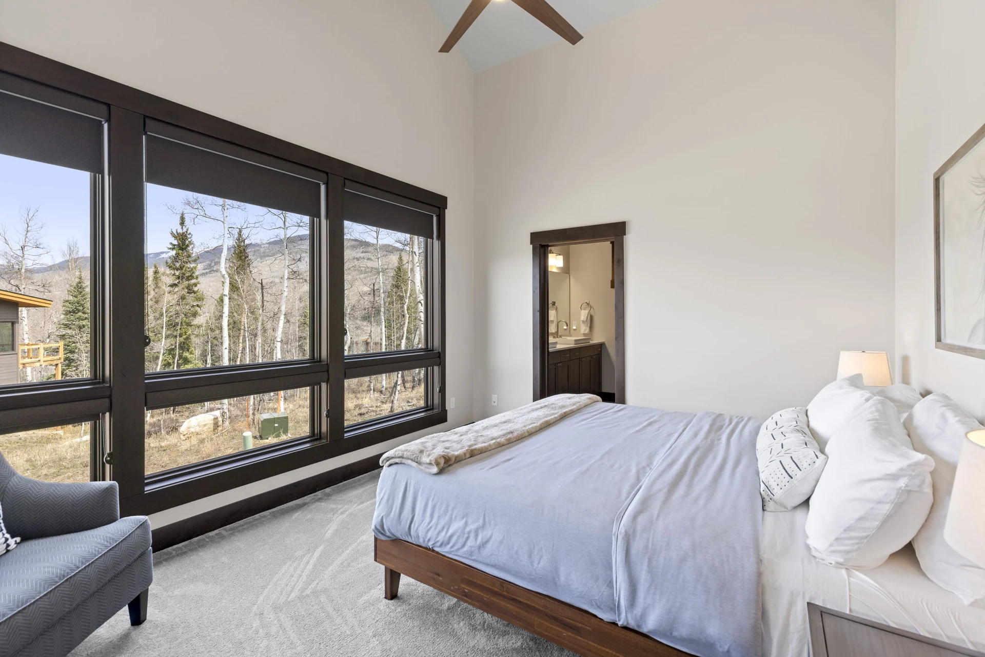 Vendette Peak, bedroom: Bedroom view toward a bank of dark-framed windows, with a gray chair and wooded hillside beyond, luxury vacation rental in Silverthorne, Colorado
