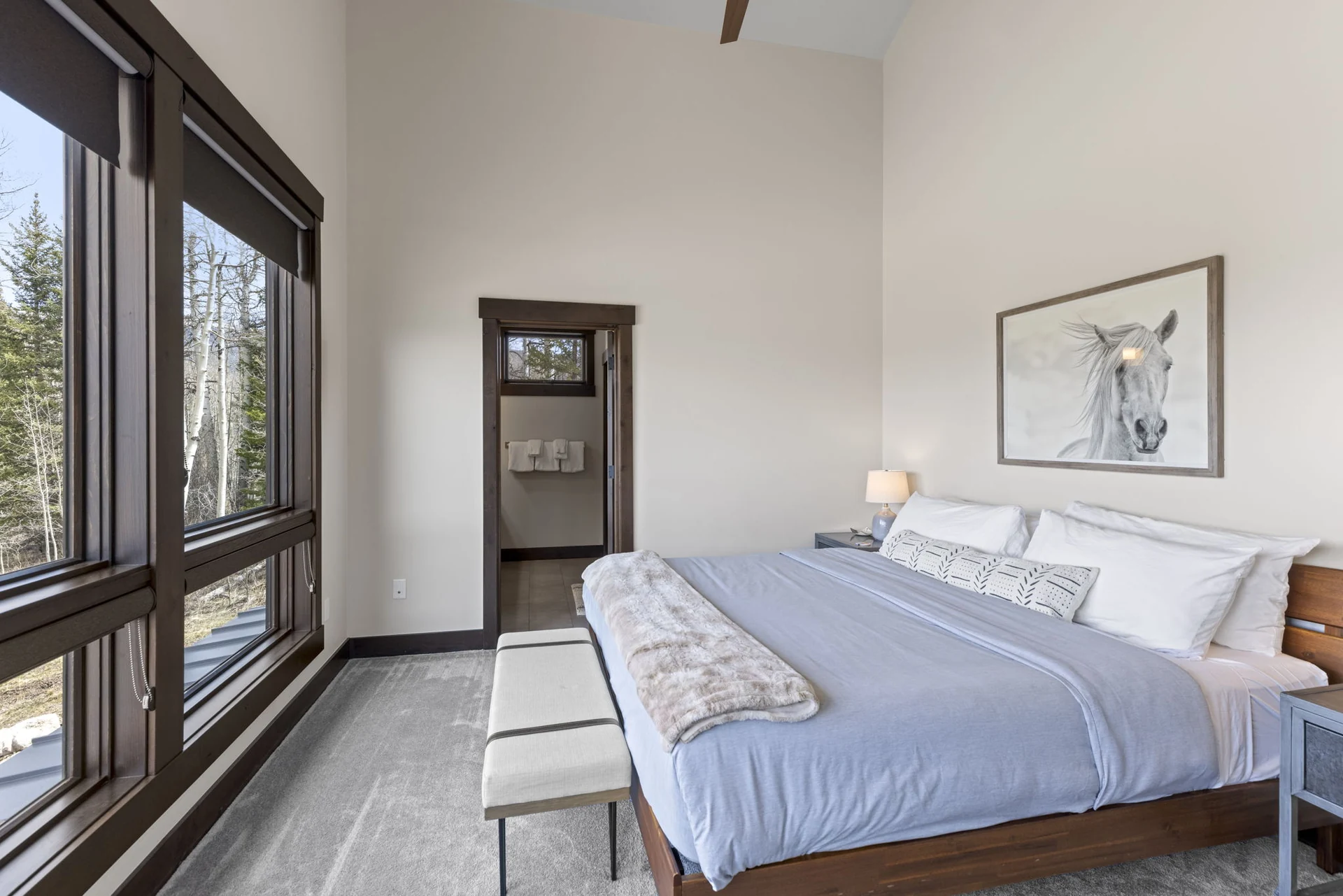 Vendette Peak, bedroom: Bed beside tall forest-facing windows, with an upholstered bench at its foot and horse artwork above, luxury vacation rental in Silverthorne, Colorado