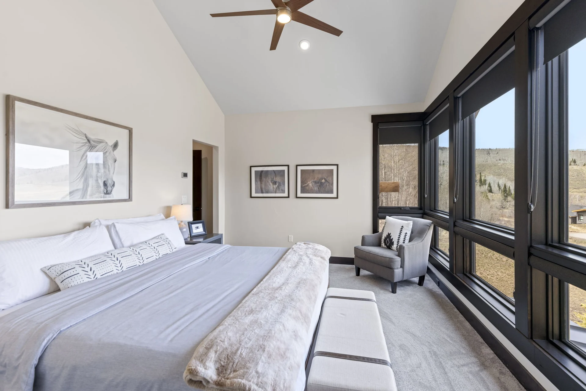 Vendette Peak, bedroom: Bright bedroom with a bed, gray chair, ceiling fan, and large corner windows overlooking the hillside, luxury vacation rental in Silverthorne, Colorado