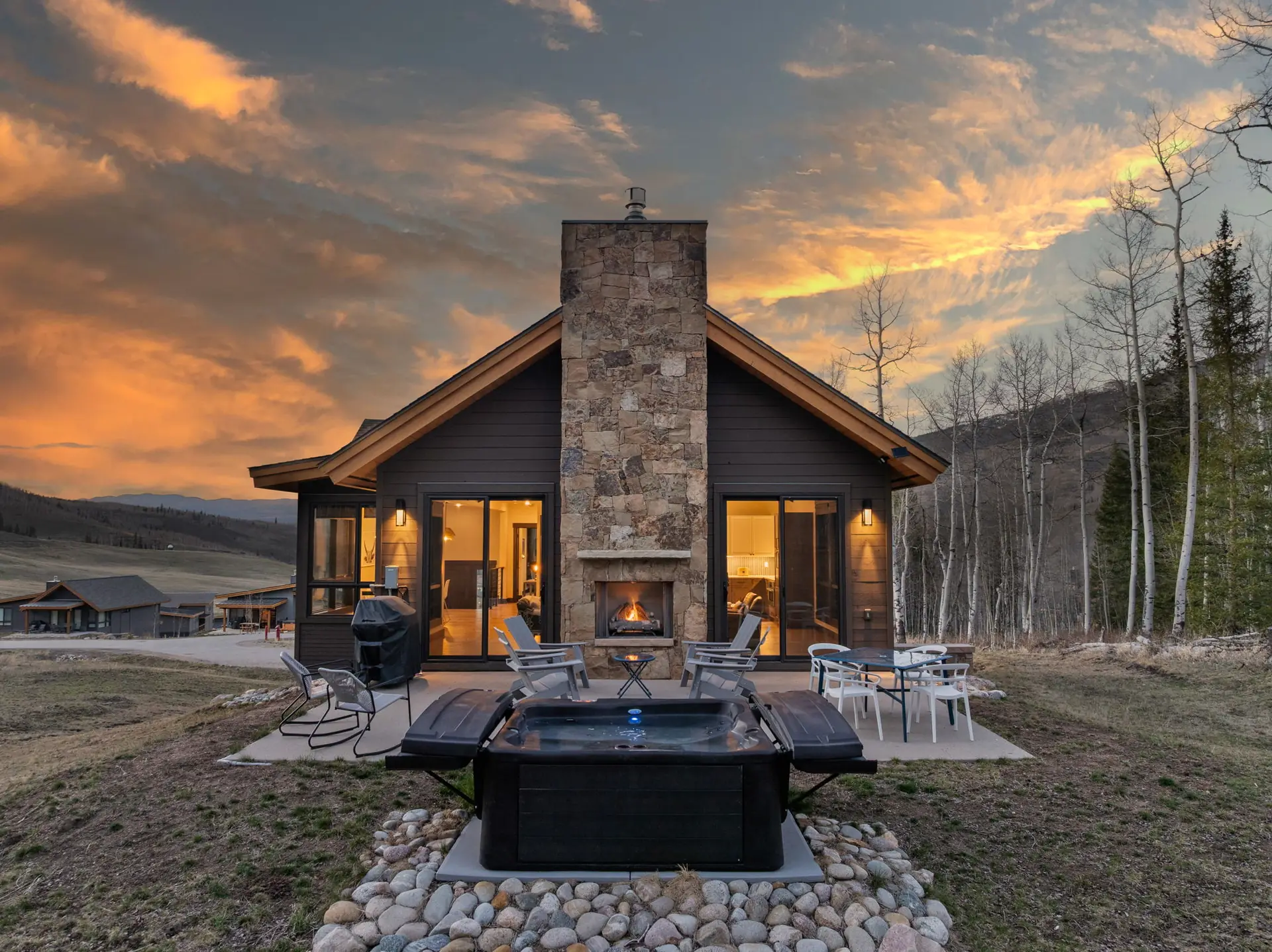 Vendette Peak, exterior: Sunset view of the home, with a stone chimney, glowing windows, patio seating, and a hot tub in front, luxury vacation rental in Silverthorne, Colorado