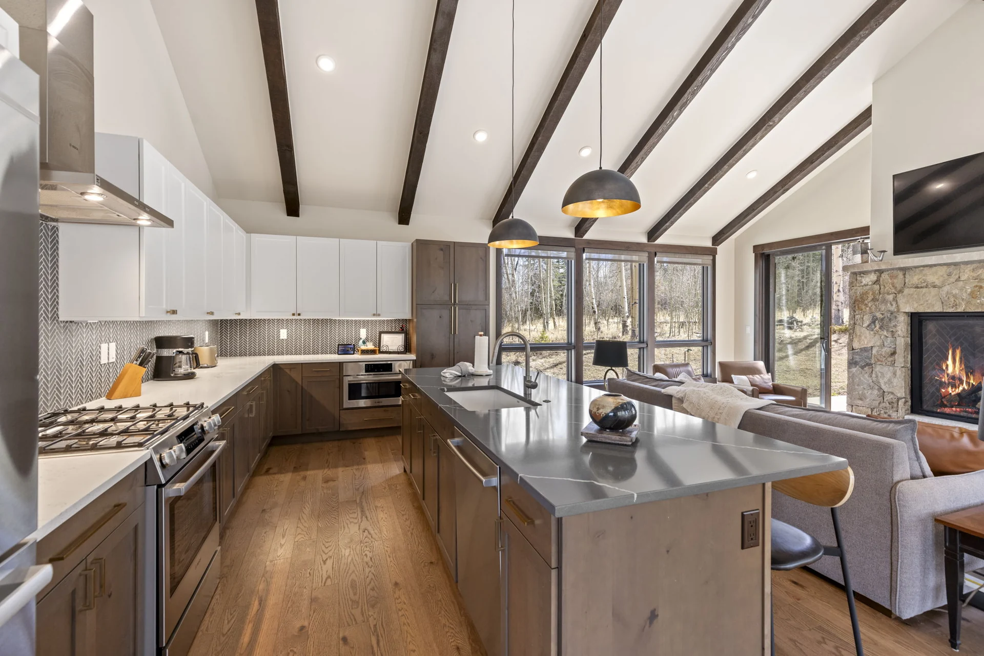Vendette Peak, kitchen: Kitchen island and cooktop beneath dark beams and pendant lights, with the living area beyond, luxury vacation rental in Silverthorne, Colorado