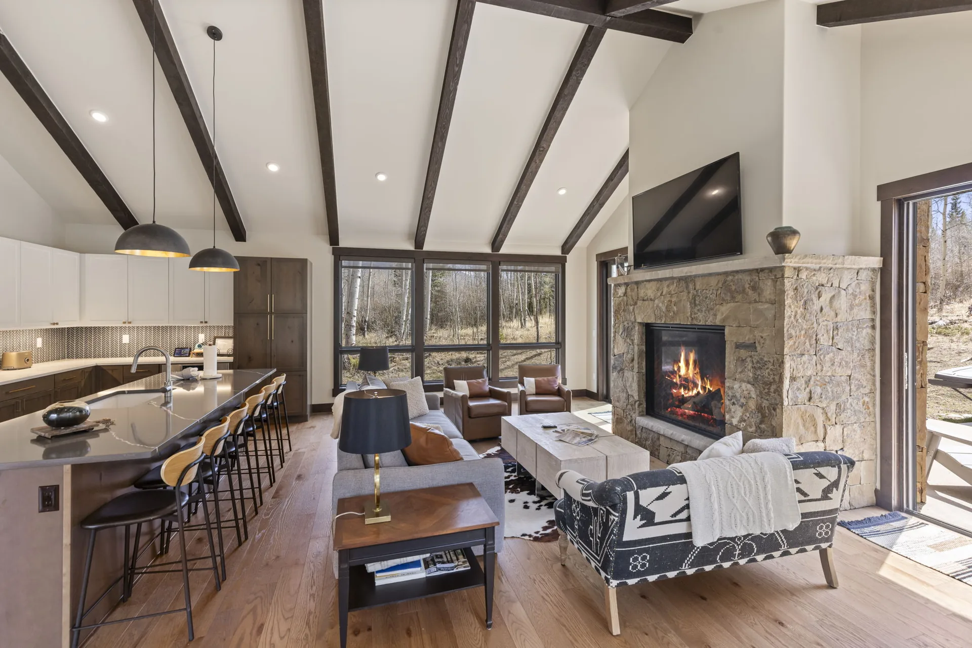 Vendette Peak, living room: Open-plan living space from the kitchen side, showing the island, sofas, stone fireplace, and window wall, luxury vacation rental in Silverthorne, Colorado